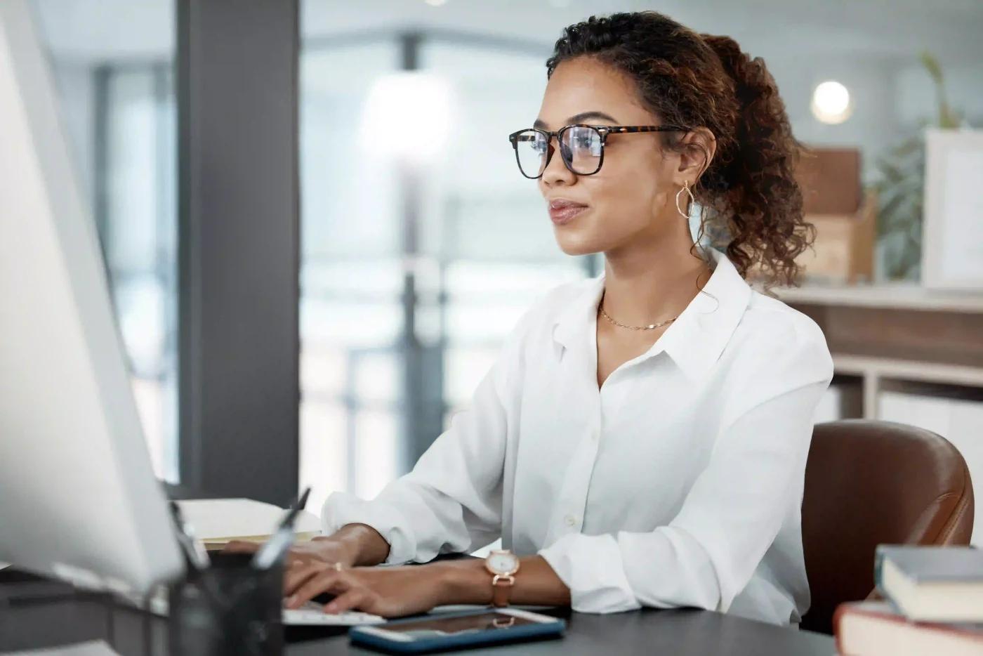 Mulher usando óculos e camisa branca trabalhando em um computador no sistema Tasklaw em um escritório moderno e iluminado.